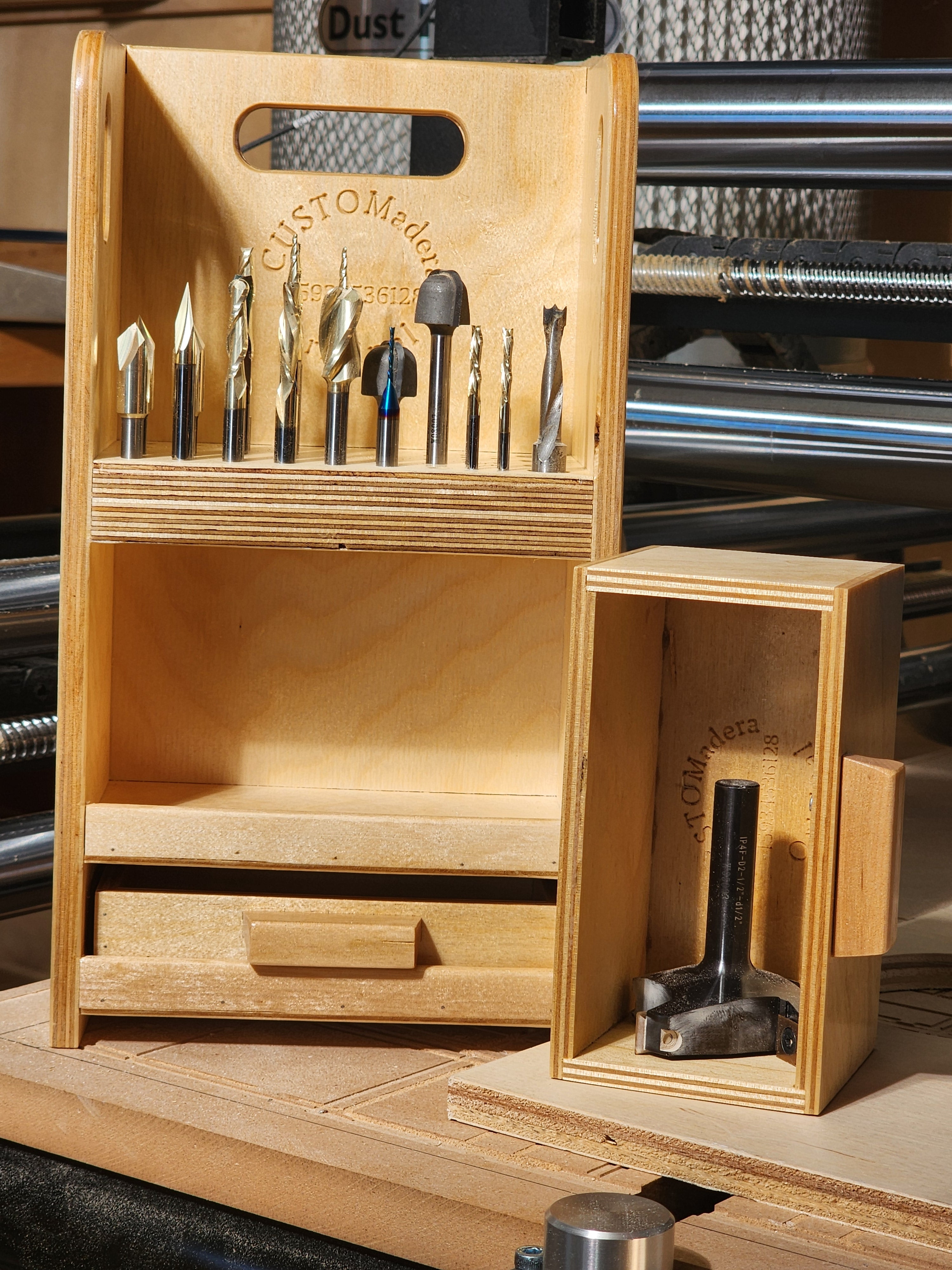 Close-up of the El Apoyo CNC™ Small CNC Bit Organizer on a CNC work surface, showing precision-drilled bit storage, the open drawer space below, integrated carry handle, CUSTOMadera branding, and a removable drawer placed beside the caddy holding a larger specialty cutter.